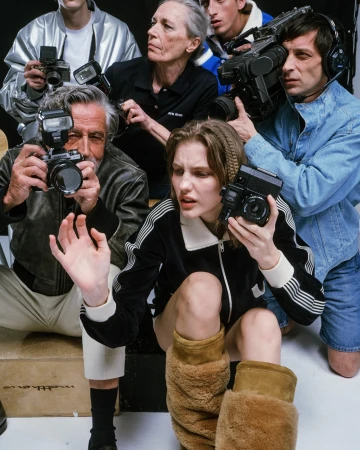 Fashion shoot featuring a model surrounded by photographers holding vintage cameras in a studio setting with a neutral background.
