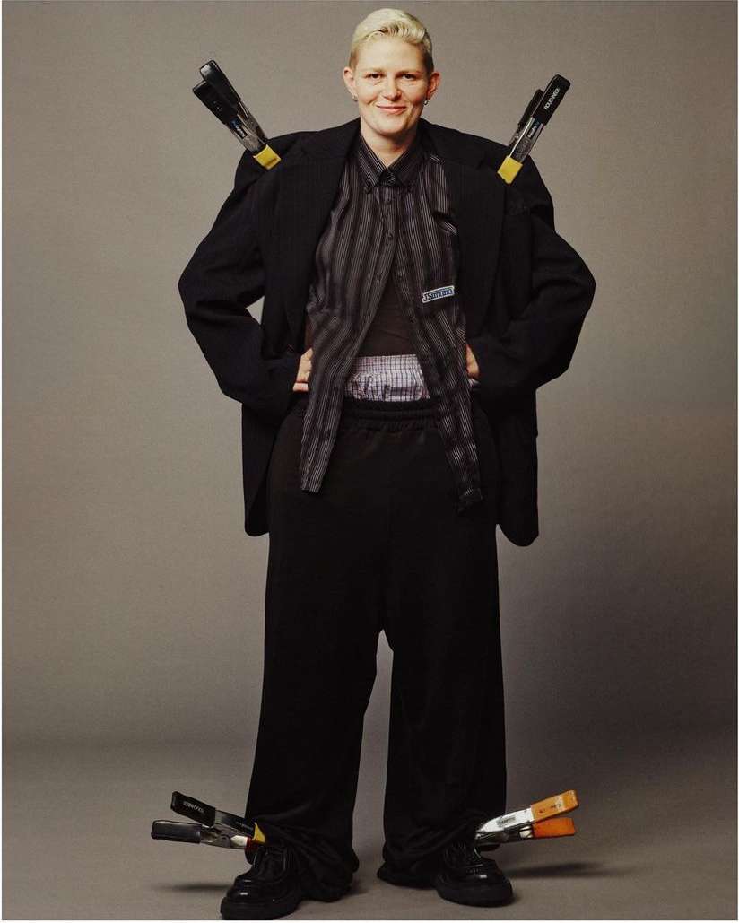 Full-length portrait of a woman with short blonde hair wearing a black blazer and trousers with clamps holding the clothing.
