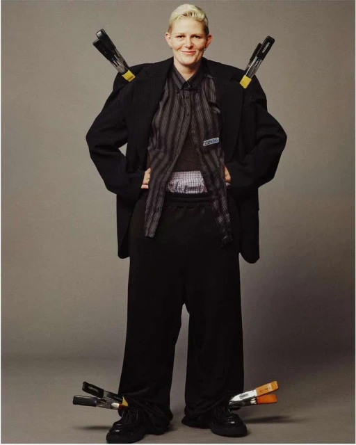 Full-length portrait of a woman with short blonde hair wearing a black blazer and trousers with clamps holding the clothing.