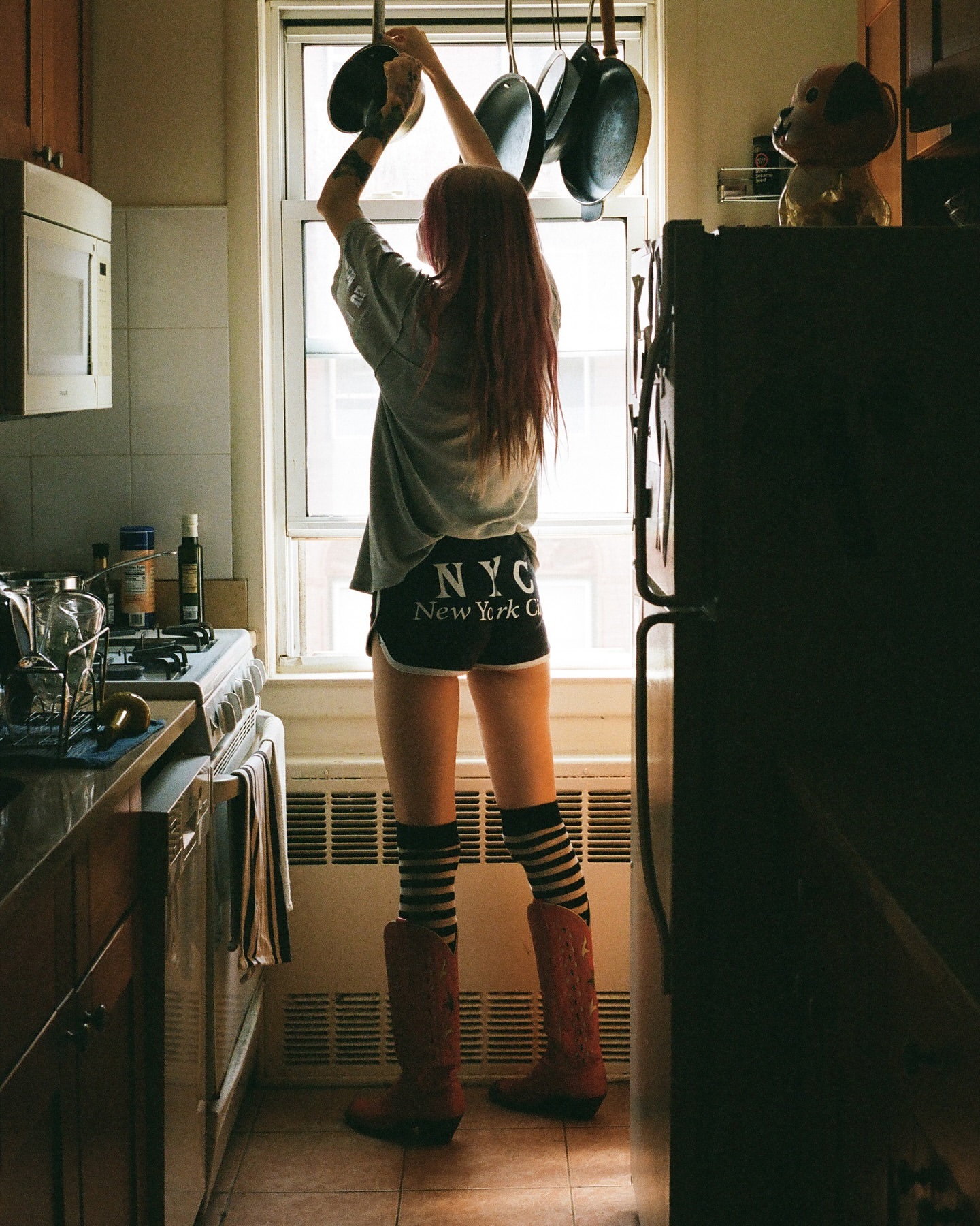 A woman in a kitchen reaching for pots and pans hanging over a window, wearing casual clothing and boots, with a view of the interior.