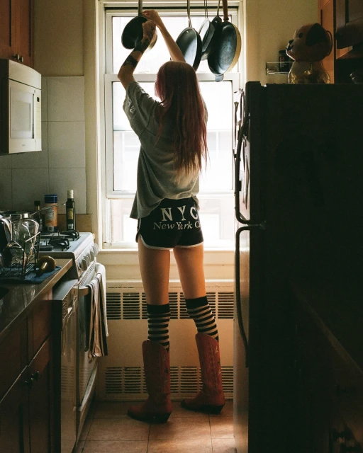 A woman in a kitchen reaching for pots and pans hanging over a window, wearing casual clothing and boots, with a view of the interior.