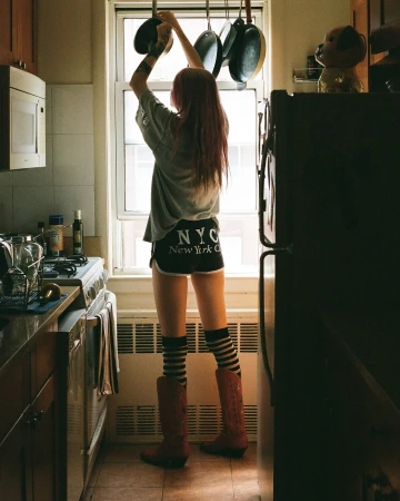 A woman in a kitchen reaching for pots and pans hanging over a window, wearing casual clothing and boots, with a view of the interior.