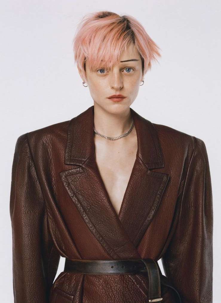 An adult female with short pink hair and a distinctive brow cut is wearing a belted brown leather jacket against a white backdrop.