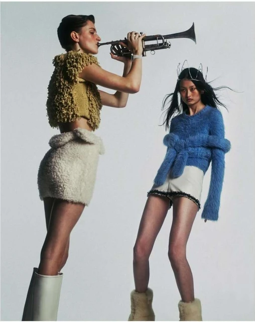Two women in stylish attire, one playing a trumpet, stand against a white backdrop in a fashion-forward studio shot.