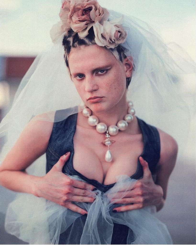 Woman in denim top and pearl necklace with flowers in her hair and veil. She is holding the top with her hands and looking at the camera.