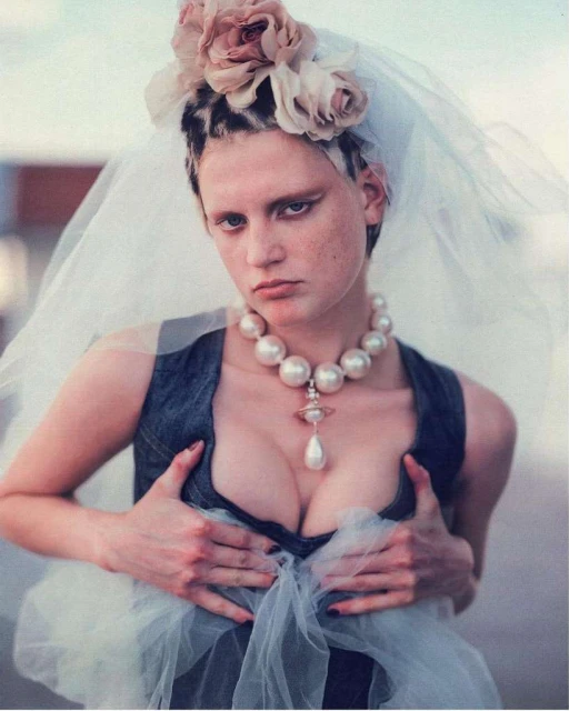 Woman in denim top and pearl necklace with flowers in her hair and veil. She is holding the top with her hands and looking at the camera.