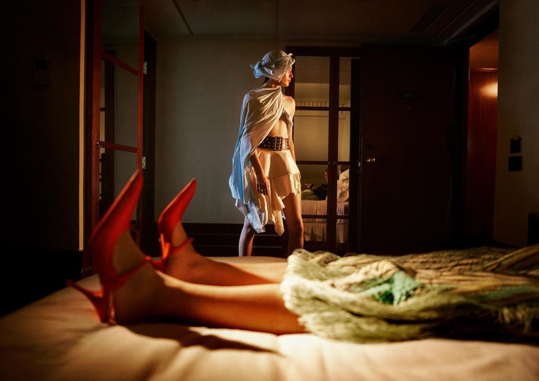 Close-up of red high heels resting on a bed with a person standing in the background of a dimly lit hotel room.