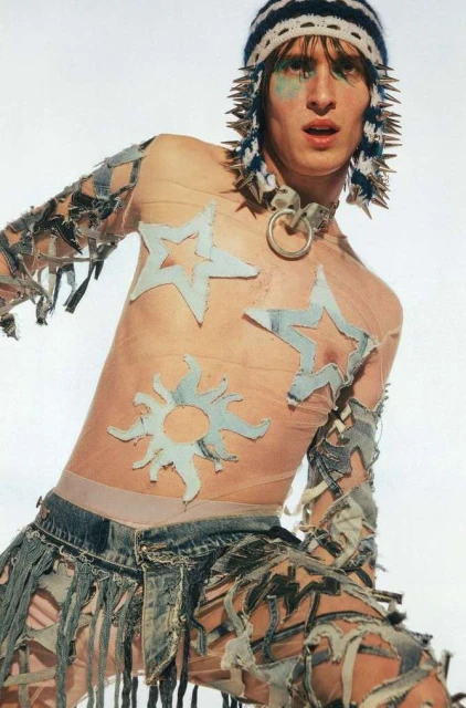 A fair-skinned adult male poses in a light studio wearing avant-garde fashion, including denim details, a striped knit hat, and bold eye makeup.
