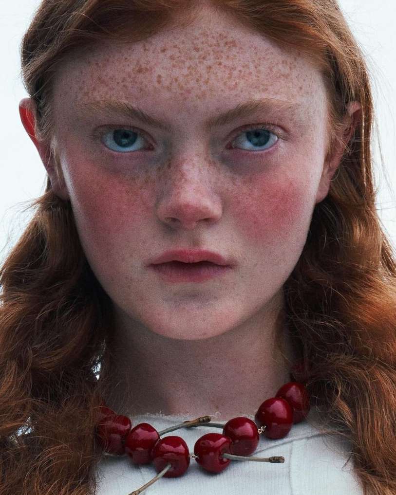 Close-up portrait of a fair-skinned young woman with red hair and freckles wearing a cherry necklace against a bright white background.