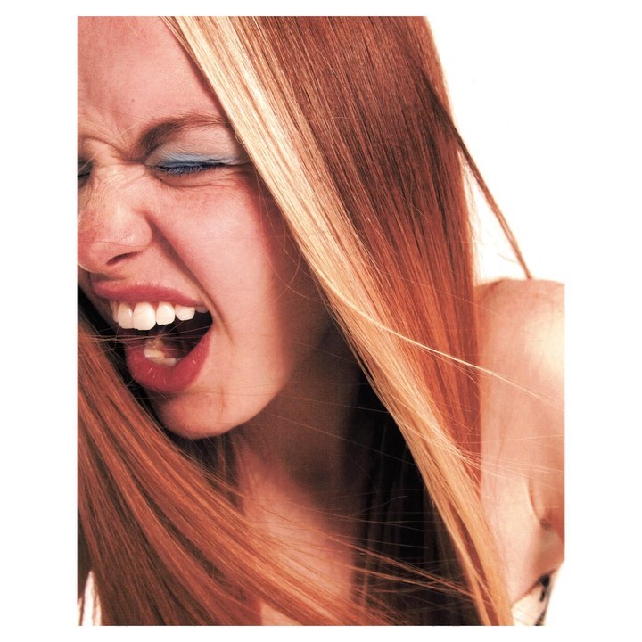 Close-up of a blonde woman with blue eyeshadow, mouth open wide in a screaming expression, with a white background, studio lighting.