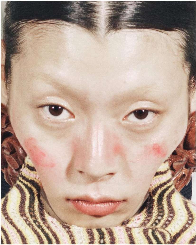 Extreme close-up of an Asian woman with pink blush, a striped turtleneck, and large wooden earrings, creating a vibrant, beauty-focused image.