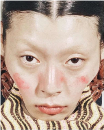 Extreme close-up of an Asian woman with pink blush, a striped turtleneck, and large wooden earrings, creating a vibrant, beauty-focused image.