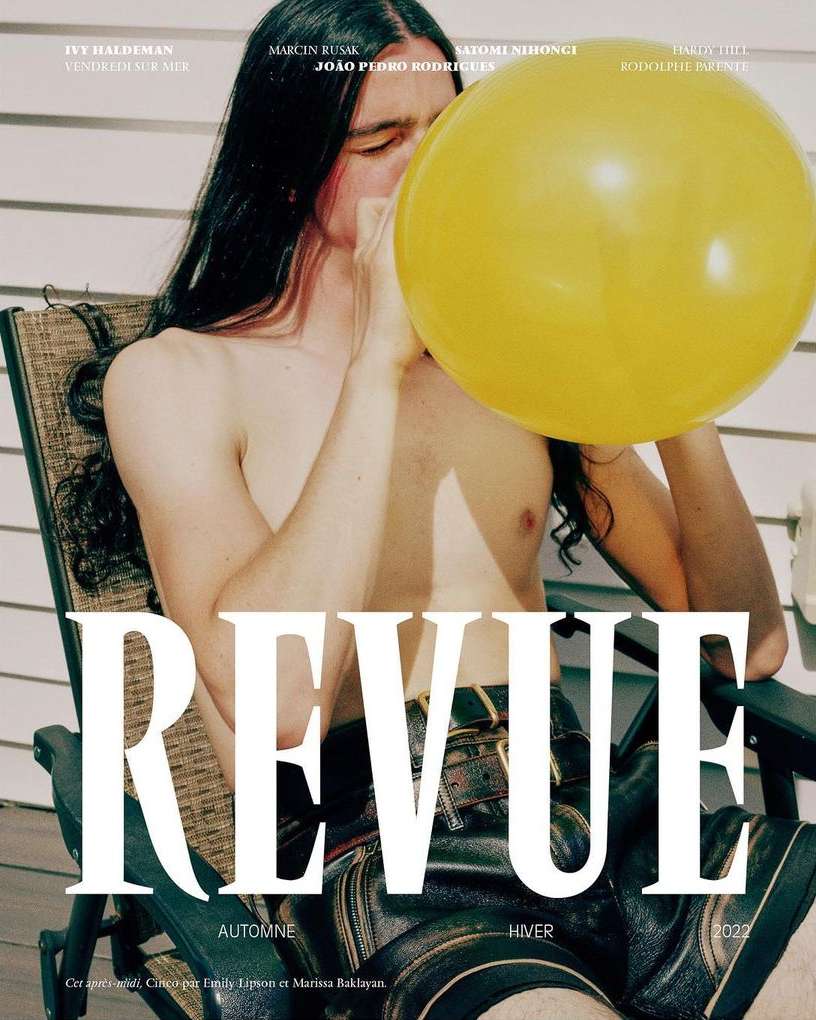 Topless person with long dark hair is seated outdoors blowing up a large yellow balloon against a backdrop of siding and furniture.