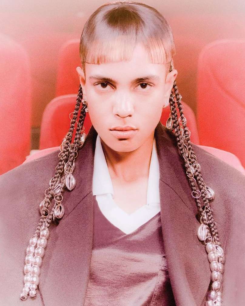 Portrait of an individual with blunt bangs and embellished braids wearing a plum jacket, styled in front of a red seating background.