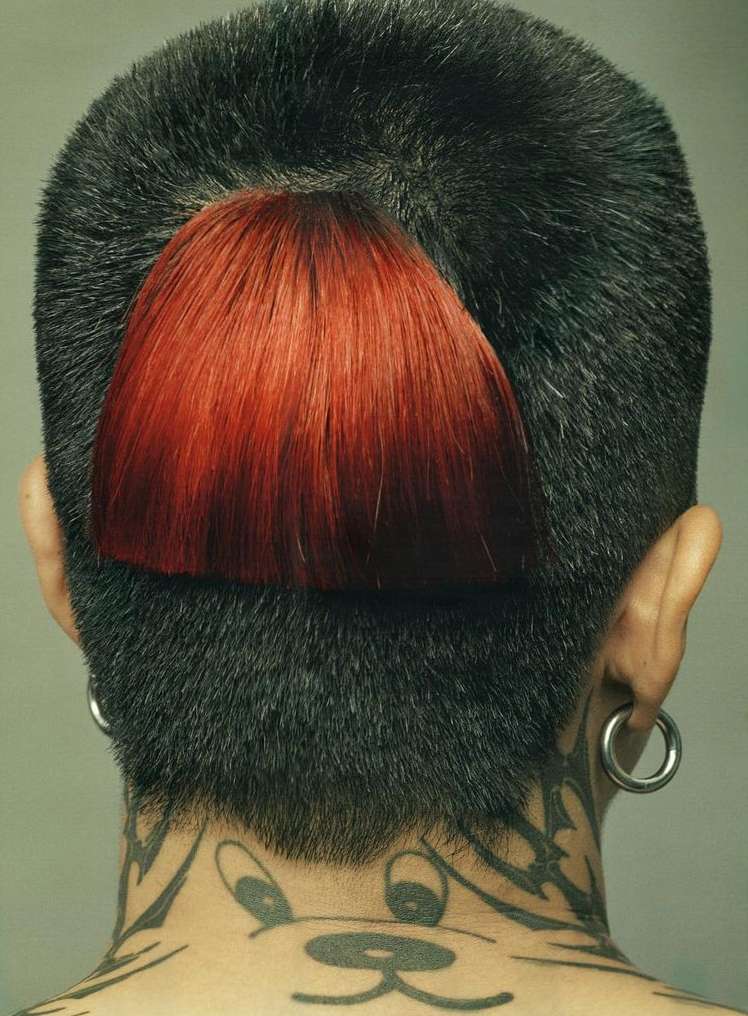 Back view of person with short black haircut, an undercut section with red hair, visible ear piercings, and tribal-style neck tattoo.