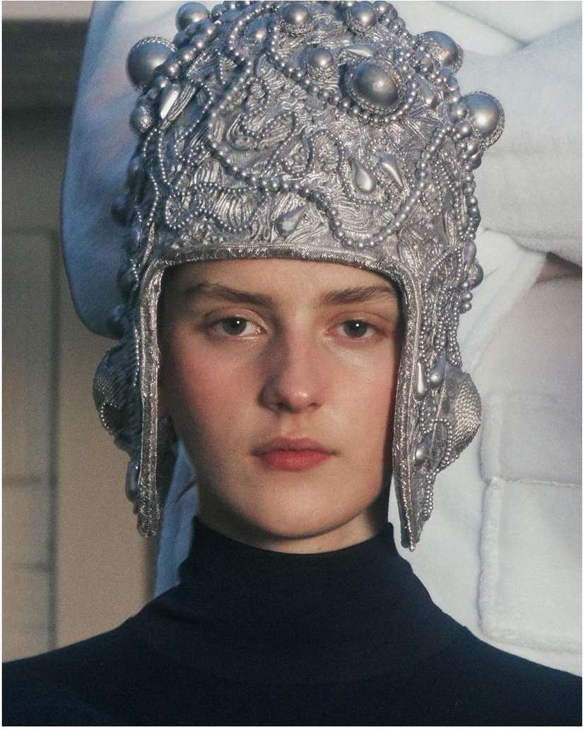Portrait of a fair-skinned woman wearing an elaborate, metallic silver-coloured headdress and a dark turtleneck shirt against a neutral backdrop.