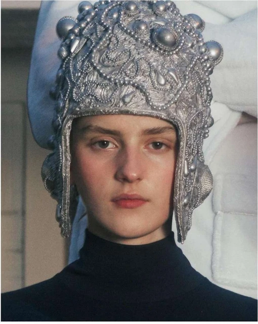 Portrait of a fair-skinned woman wearing an elaborate, metallic silver-coloured headdress and a dark turtleneck shirt against a neutral backdrop.