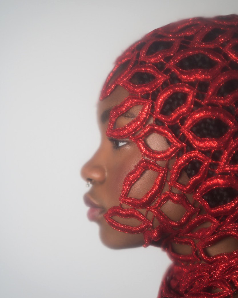 Close-up profile of a Black woman wearing a red lace head covering in a studio setting with soft, diffused lighting against a white backdrop.
