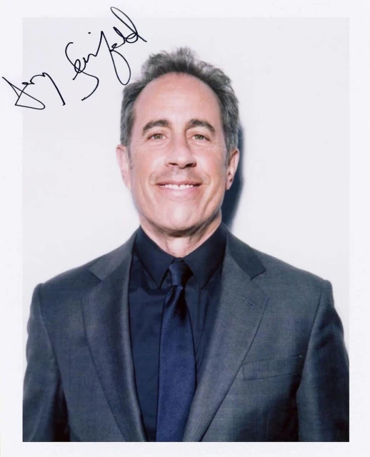 Portrait of Jerry Seinfeld smiling in a grey suit, dark shirt, and tie, with his signature visible in the top left corner. 