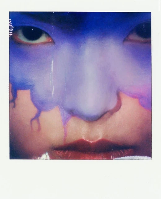 Close-up shot of a person's face with blue and purple paint-like makeup on the forehead, nose, and cheeks; dark red lipstick.