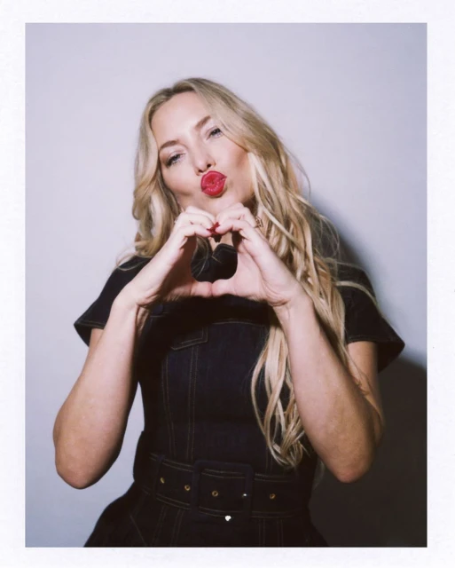 Blonde woman in dark denim dress making heart shape with hands and blowing kiss, posing against a light background, medium shot.