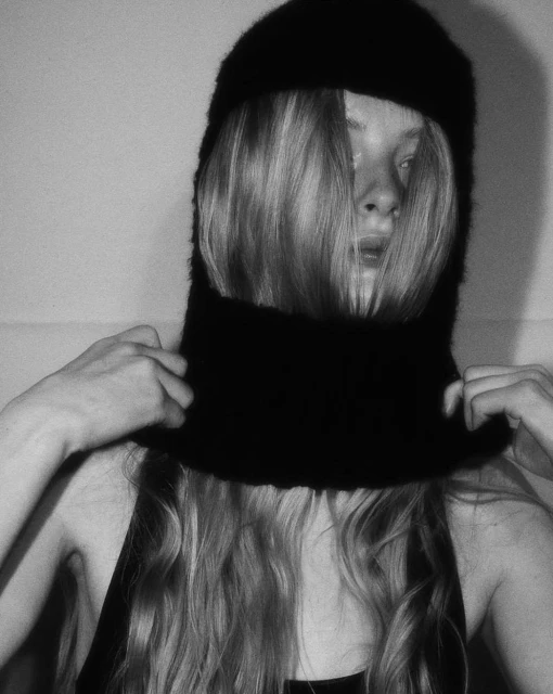 A woman with long blonde hair adjusts a black balaclava in a close-up, black-and-white studio portrait with soft, diffused lighting.