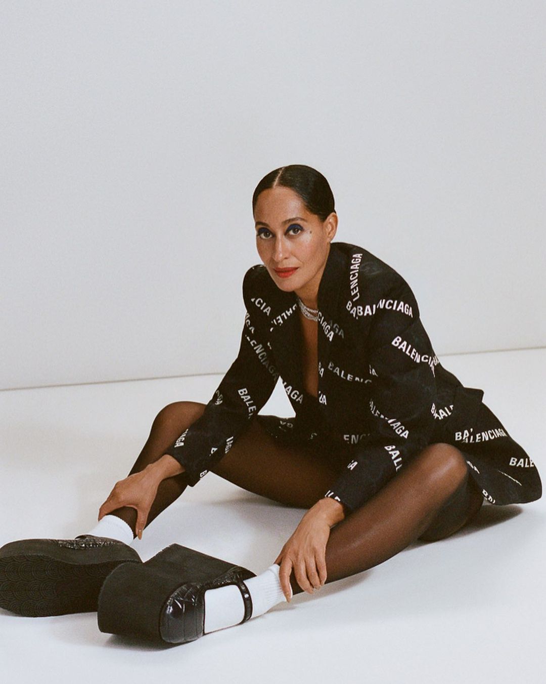 Adult female poses indoors, wearing a black and white Balenciaga blazer, black tights, white socks, and black platform Mary Jane shoes.