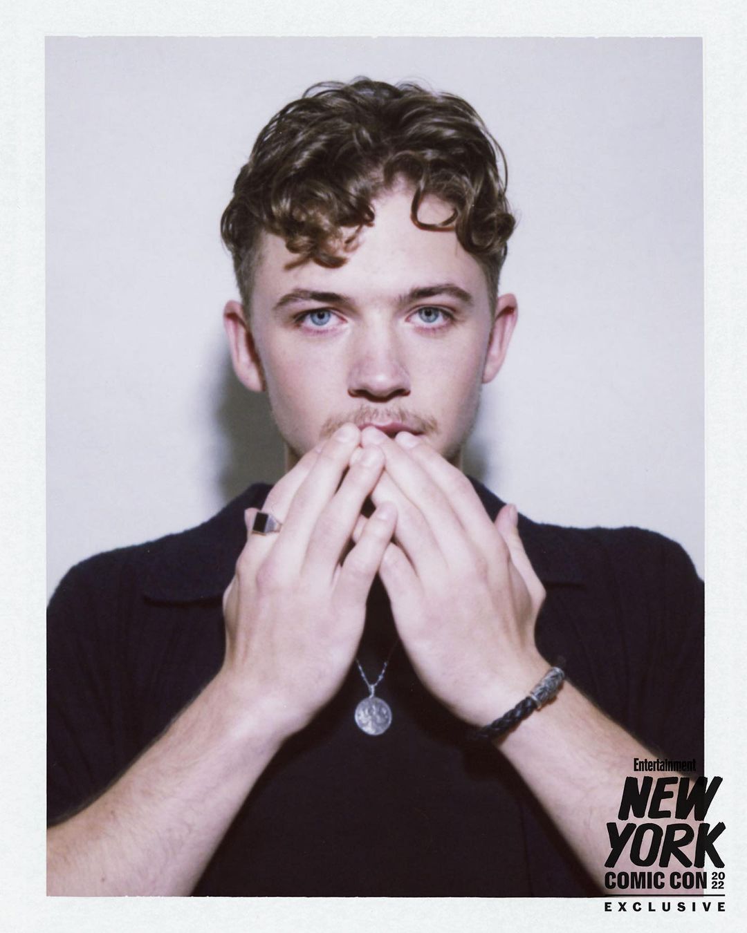Headshot of a fair-skinned young adult male with curly brown hair, blue eyes, and light stubble, wearing a black collared shirt.