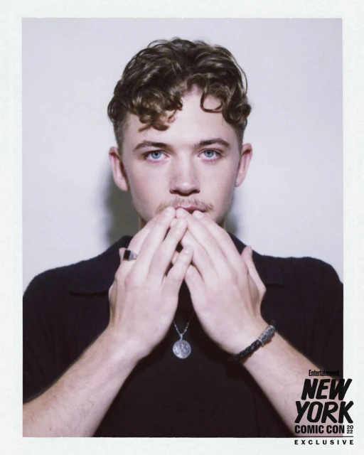 Headshot of a fair-skinned young adult male with curly brown hair, blue eyes, and light stubble, wearing a black collared shirt.