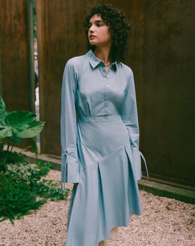 A stylish woman with curly hair in a light blue dress stands in an outdoor setting with gravel and greenery, creating a fashion-forward visual.