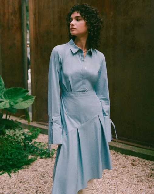 A stylish woman with curly hair in a light blue dress stands in an outdoor setting with gravel and greenery, creating a fashion-forward visual.