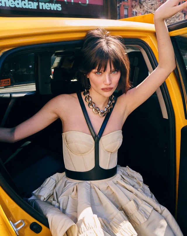 A woman in a beige dress and black harness poses in a yellow taxi, looking directly at the camera with city buildings in the background.
