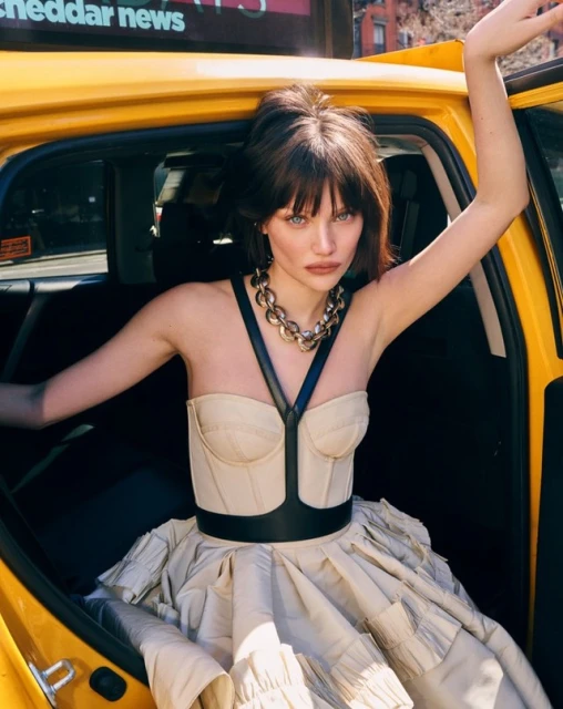 A woman in a beige dress and black harness poses in a yellow taxi, looking directly at the camera with city buildings in the background.