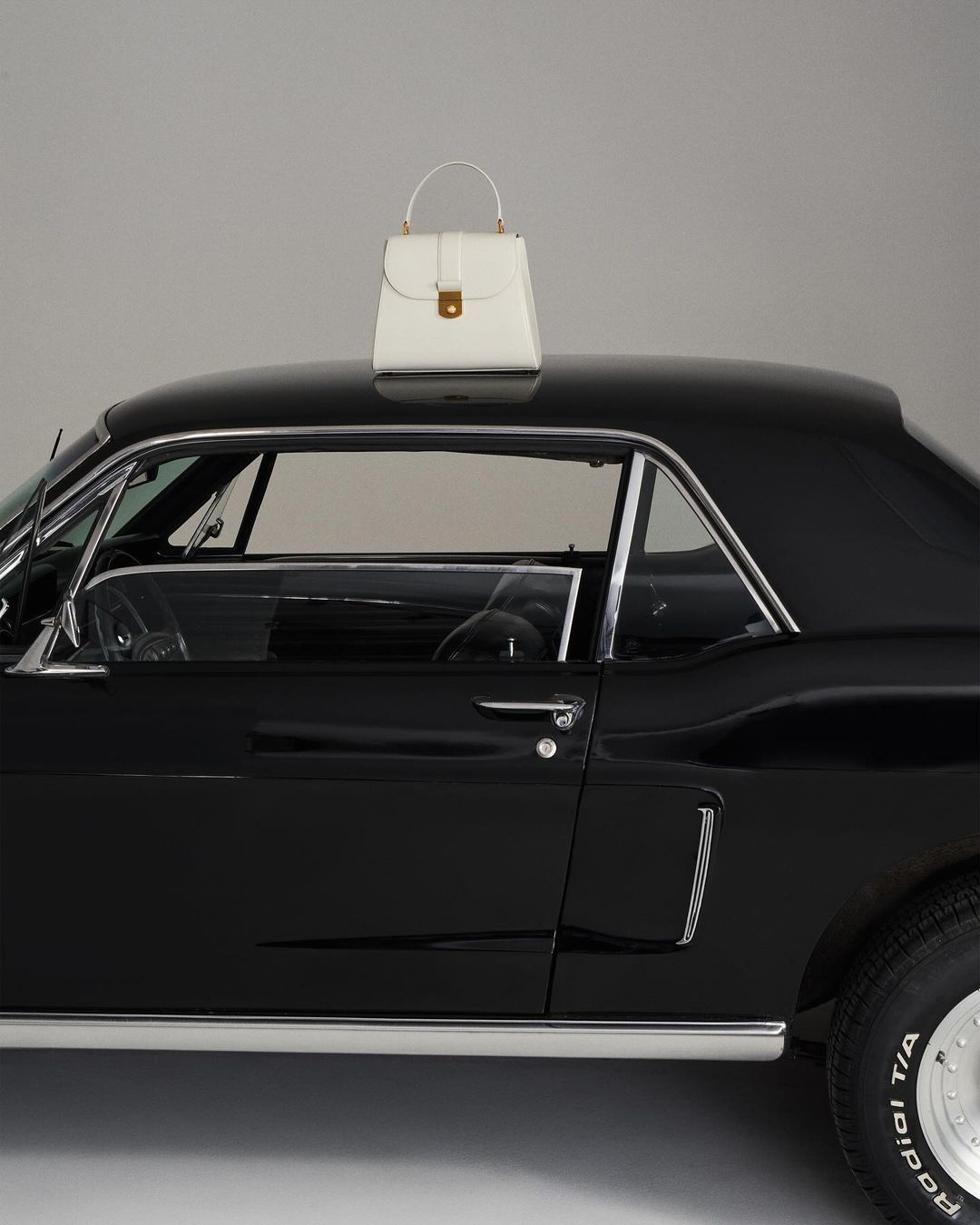 Elegant white leather handbag with top handle resting on a sleek black classic car, set against a plain grey backdrop in studio lighting.