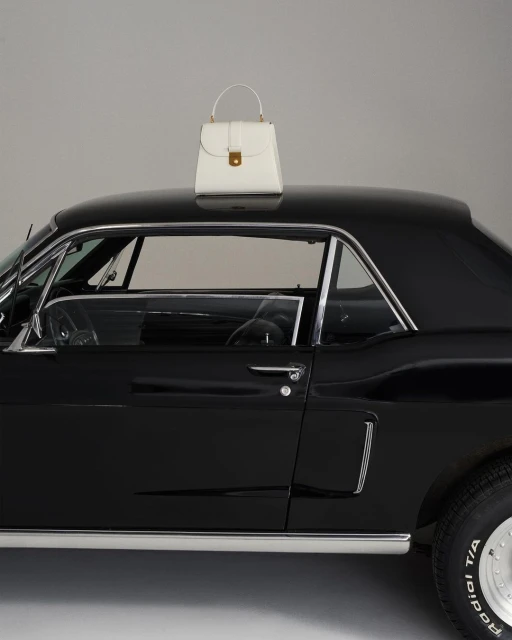 Elegant white leather handbag with top handle resting on a sleek black classic car, set against a plain grey backdrop in studio lighting.