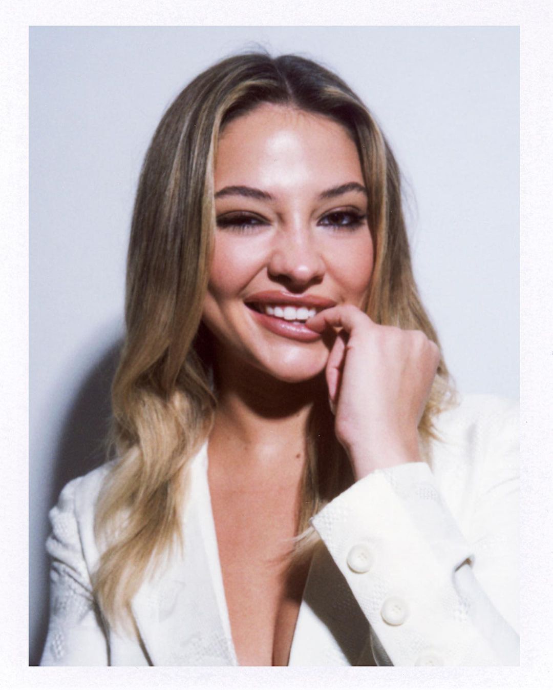 Blonde woman in white blazer smiles with hand near lips against white backdrop in a studio portrait with shallow depth of field.