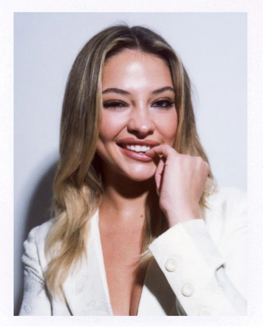 Blonde woman in white blazer smiles with hand near lips against white backdrop in a studio portrait with shallow depth of field.
