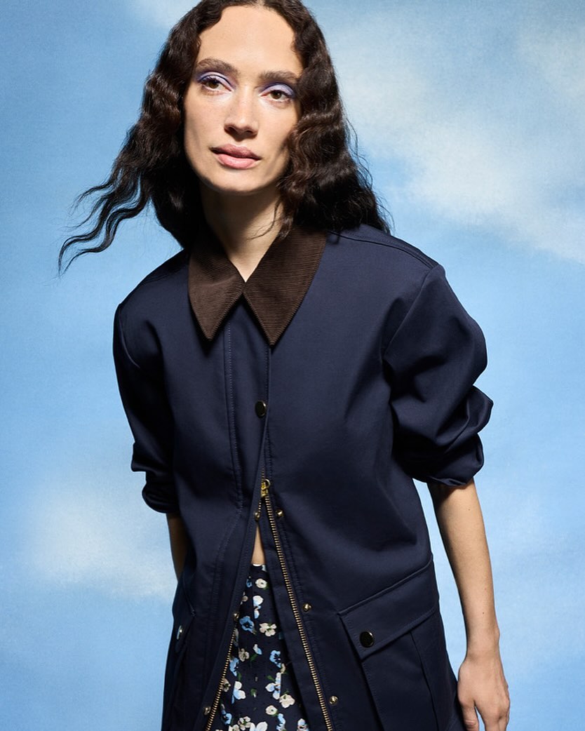Portrait of a woman with dark curly hair wearing a navy jacket with a brown collar in front of a light blue sky with clouds.