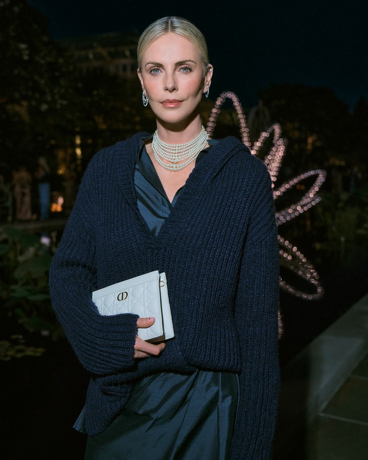 Elegant adult female in navy sweater holds designer purse at an evening event with decorative lighting, wearing a pearl necklace and earrings.