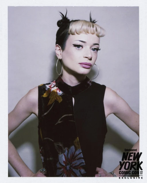 Stylish woman with black and blonde hair, hoop earrings, and a floral dress poses indoors against a neutral backdrop, New York Comic Con.