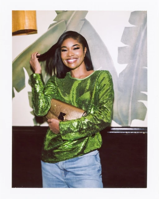 Smiling woman in a sequin top and jeans holds a gold clutch against a leaf-patterned wall, illuminated by an overhead light.