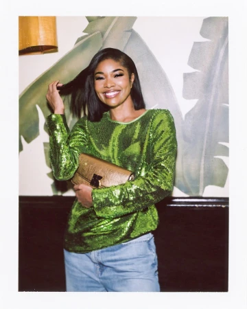 Smiling woman in a sequin top and jeans holds a gold clutch against a leaf-patterned wall, illuminated by an overhead light.