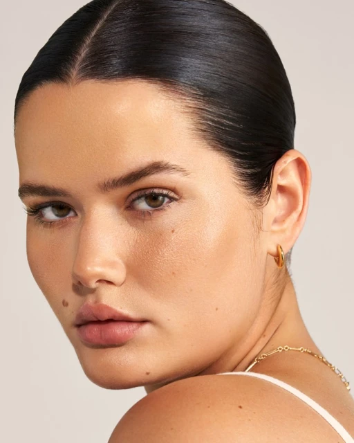 Close-up of an adult woman with dark hair and brown eyes wearing gold hoop earrings in a bright indoor studio lighting setting.