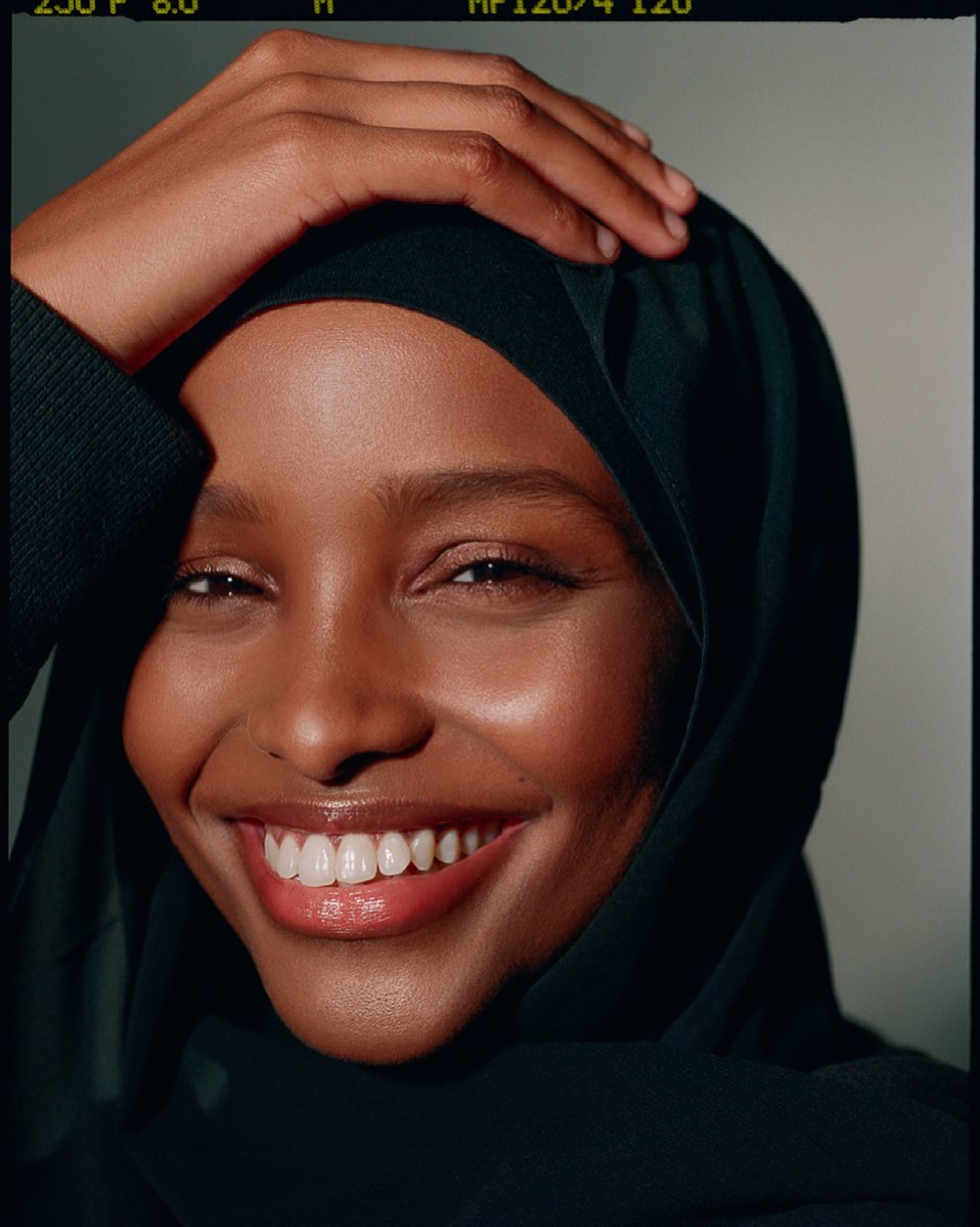 Close-up portrait of a smiling, dark-skinned adult female wearing a black hijab and a black sweater, looking directly at the camera.