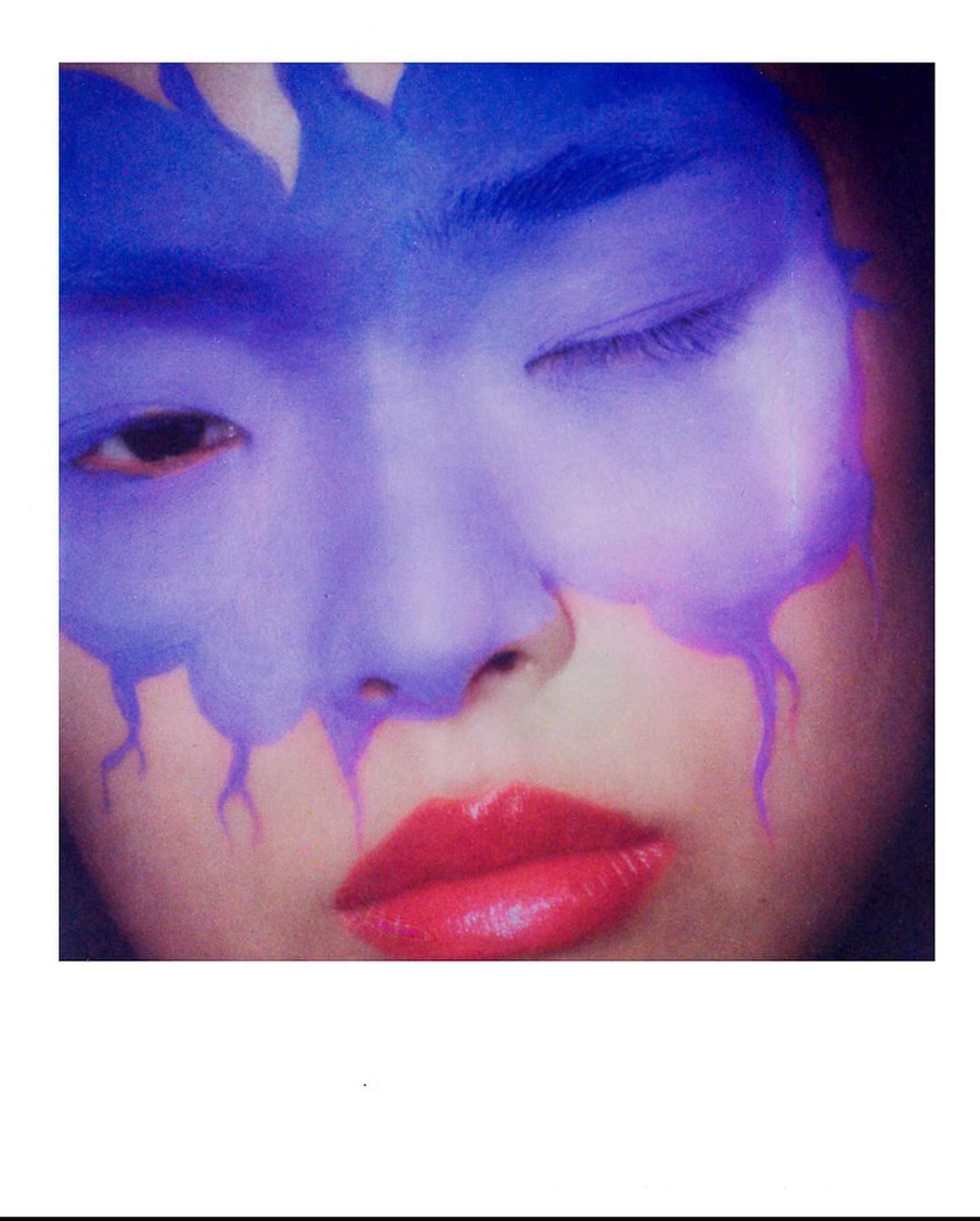 Close-up portrait of a person with blue face paint, red lips, and one eye closed, showcasing makeup artistry and beauty aesthetics.