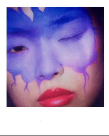 Close-up portrait of a person with blue face paint, red lips, and one eye closed, showcasing makeup artistry and beauty aesthetics.
