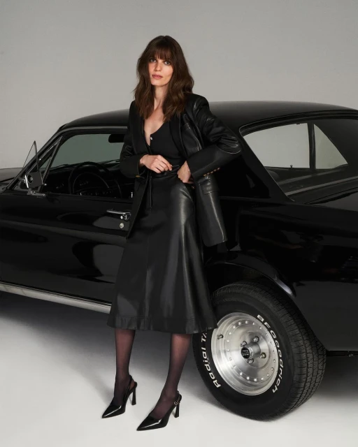 Woman in black leather outfit poses next to a black classic car in a studio setting, showcasing fashion and automotive style.