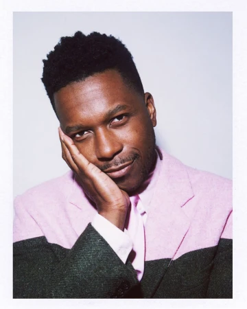 Close-up studio portrait of adult male with hand on cheek, wearing color-block blazer. Soft, diffused lighting emphasizes facial features.