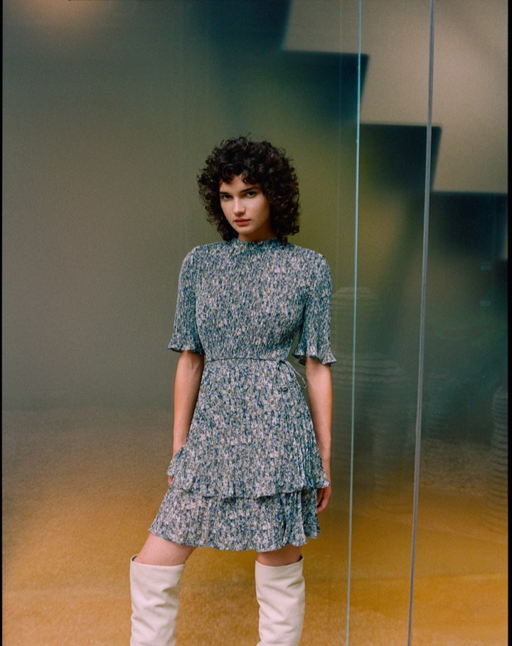 Model with curly dark hair wearing a patterned midi dress and knee-high boots in a contemporary interior.
