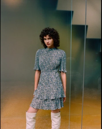 Model with curly dark hair wearing a patterned midi dress and knee-high boots in a contemporary interior.